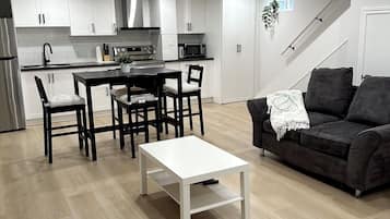 A modern kitchen with white cabinets, a dining area with a white table and chairs, and a dark grey sofa in a living room.