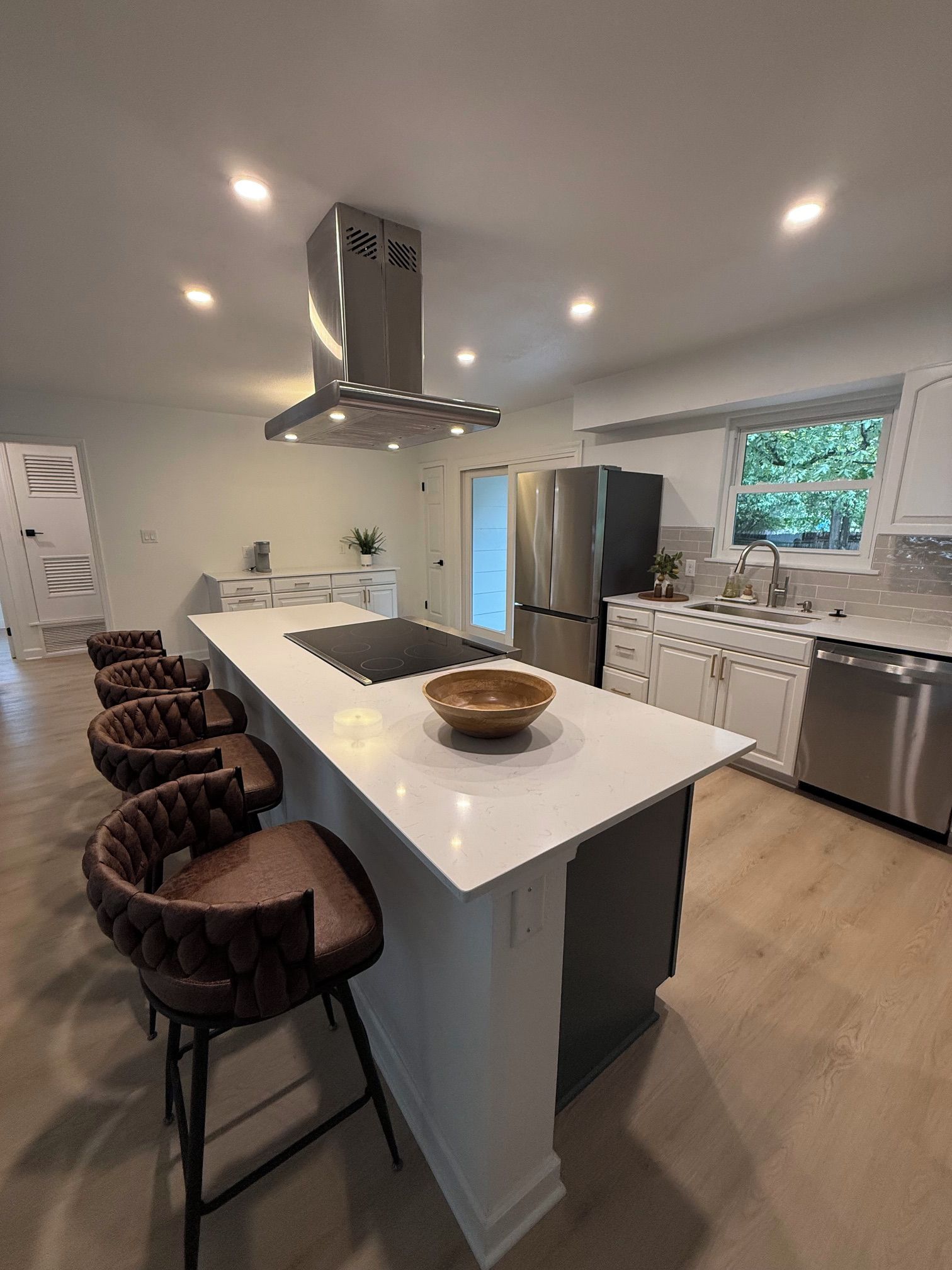 A modern kitchen with a central island, bar stools, stainless steel appliances, and a view of greenery outside.