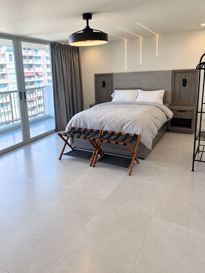 Ein modernes Schlafzimmer mit einem großen Bett, einer Bank, einem Nachttisch und Blick auf die Stadt durch ein großes Fenster.