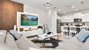 Ein modernes Wohnzimmer mit Flachbildfernseher, einer Couch, einem Couchtisch und einer Küchenzeile mit weißen Schränken und Geräten.