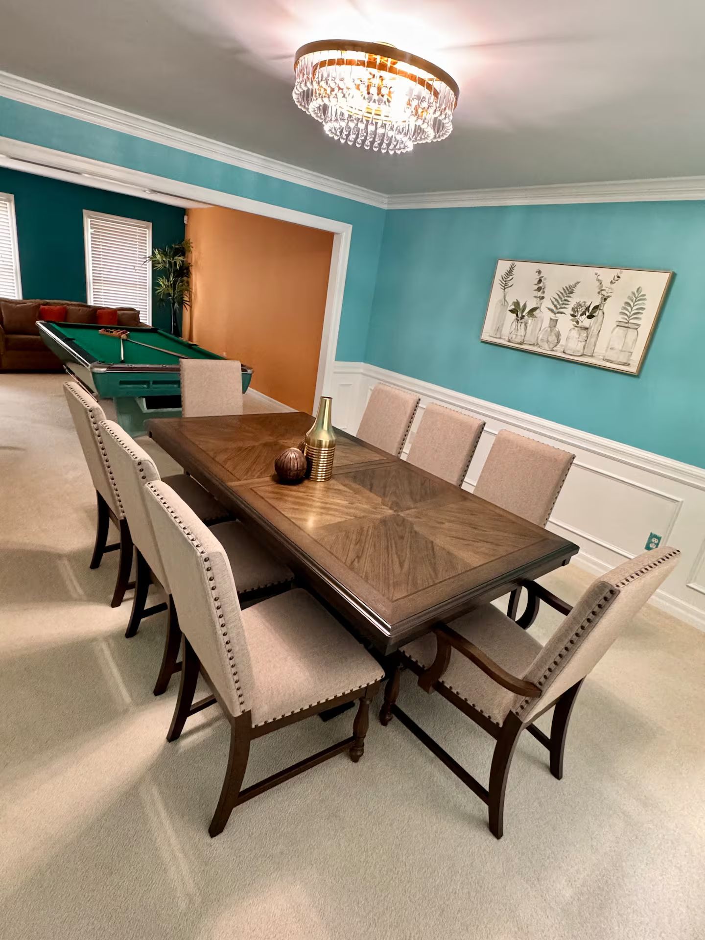 Una sala da pranzo con tavolo in legno, sedie imbottite beige, un lampadario, un tavolo da biliardo e una parete con un dipinto a soggetto botanico.
