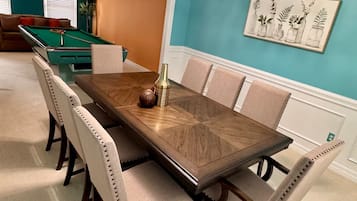 Una sala da pranzo con tavolo in legno, sedie imbottite beige, un lampadario, un tavolo da biliardo e una parete con un dipinto a soggetto botanico.