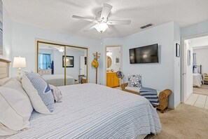 A bedroom with a bed, a ceiling fan, a television, a mirror, and a door leading to another room. - Ground-Floor Sun Beach Club, 2 Kings, Pool and Hot Tub, Walk to Car-Free Beach (New Smyrna Beach)