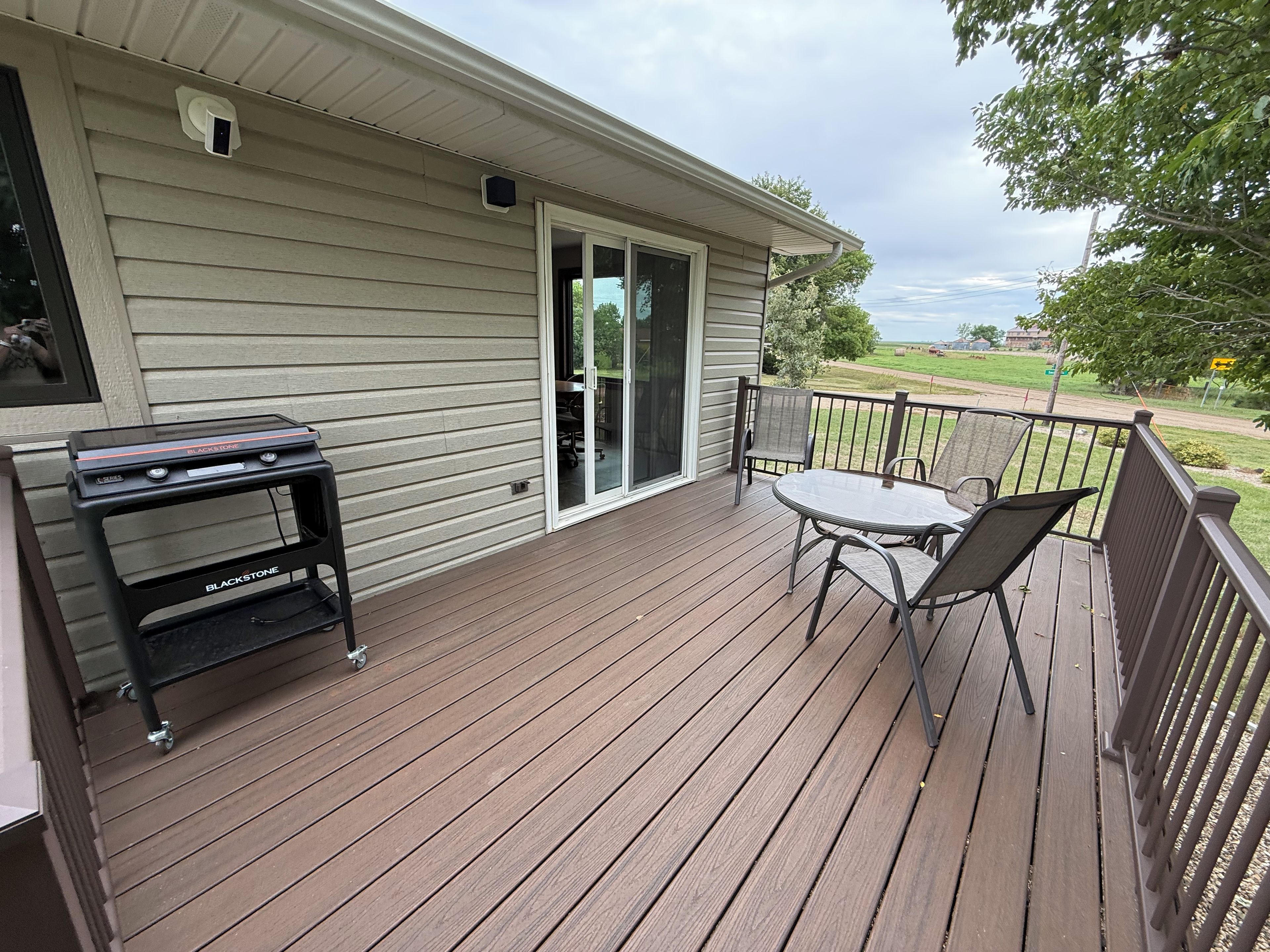 Um deck de madeira com churrasqueira e mesa com cadeiras, uma porta de correr e vista para o exterior.