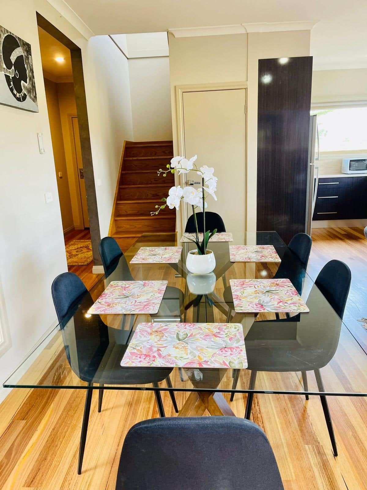 Uma área de jantar com mesa de vidro, rodeada por cadeiras, um vaso com flores brancas e uma escadaria ao fundo.