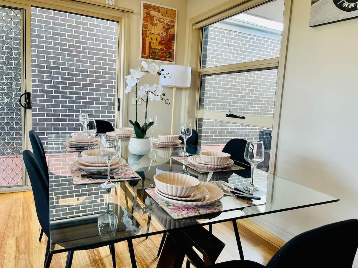A dining area with a glass table, white plates, and a vase of white flowers.