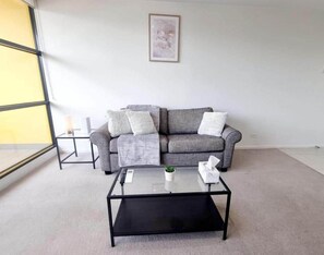 A living room with a grey sofa, a glass coffee table, and a window on the left. - Skyline Views 1BR Opp Crown w/Free Parking (Southbank)