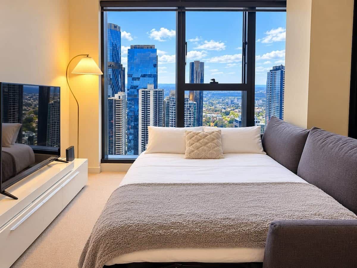 A bedroom with a large bed, a TV on a stand, a lamp, and a city view through large windows.