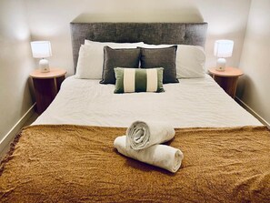 Cama bem arrumada com manta marrom texturizada e toalhas brancas dobradas, duas luminárias de cabeceira e um encosto de cama.