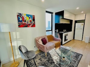 Uma sala de estar moderna com sofá, poltrona, mesa de centro de vidro, pintura na parede e área de cozinha com armários e eletrodomésticos.