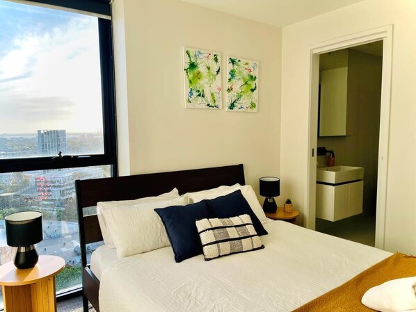 Quarto de hotel com uma cama grande, duas mesas de cabeceira, uma janela com vista para a cidade e uma porta que dá para um banheiro.