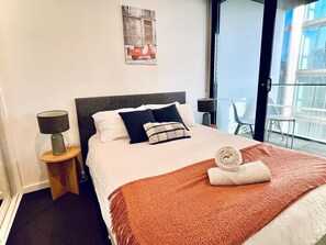 Quarto de hotel com cama, mesa de cabeceira com luminária, janela com vista para prédios e uma cadeira perto da janela.