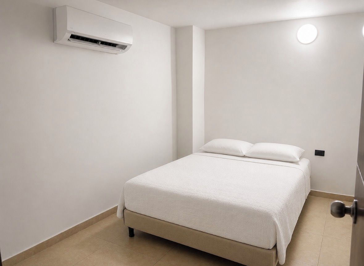 A single bed with a white mattress and pillows, a wall-mounted air conditioner, and a door with a round light fixture.