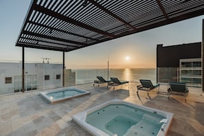 A rooftop terrace with two hot tubs and lounge chairs, overlooking a body of water during sunset. - Spacious, modern studio apartment 80 meters from Bello Horizonte Beach with an ocean view (Unknown)