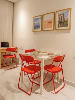 A dining area with a white table and red chairs, a laptop on a desk, and three framed pictures on the wall. - Spacious, modern studio apartment 80 meters from Bello Horizonte Beach with an ocean view (Unknown)