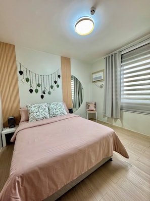 Ein Schlafzimmer mit Bett, Nachttisch, Lampe, Wanddekoration, Fenster mit Jalousien und Deckenleuchte.
