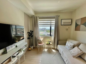 A living room with a television, a sofa, a chair, a small table with a plant, and a window with curtains. - Cozy Staycation • Free Parking • 300Mbps WiFi • Taytay, Rizal (Taytay)