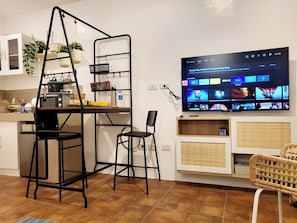 A modern living room with a TV displaying streaming services, a kitchen area with a ladder and shelves, and a wicker chair. - Cozy La Bella Tagaytay Stay Pool View + Work Space (Tagaytay City)