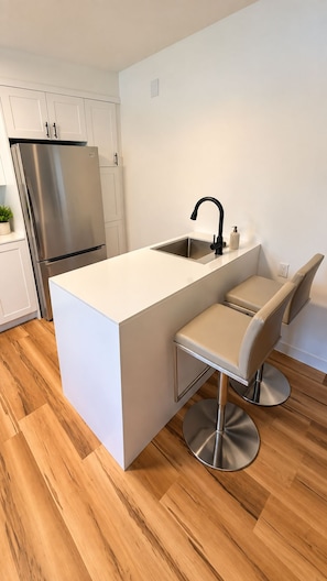 A modern kitchen with a stainless steel refrigerator, a white countertop island with a sink, and a bar stool. - Vintage & lumineux • Secteur calme (Montréal)