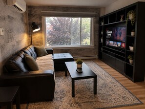 Ein modernes Wohnzimmer mit einer Ledersofa, einem Couchtisch mit Pflanze, einem großen Fenster mit Blick auf Bäume und einer schwarzen Unterhaltungsanlage mit einem Fernseher, der 'Stranger Things' zeigt.