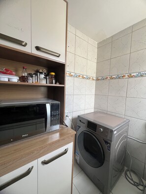 A compact kitchen with a microwave, a washing machine, and a shelf with kitchen utensils. - 2-Bedroom Apartment in the Central Area of ​​São Luís, MA. (São Luís)