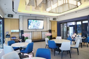A modern hotel lobby with round tables, blue chairs, a large TV screen, and a decorative plant. - Mansionite Hotel Kudus (Kudus)