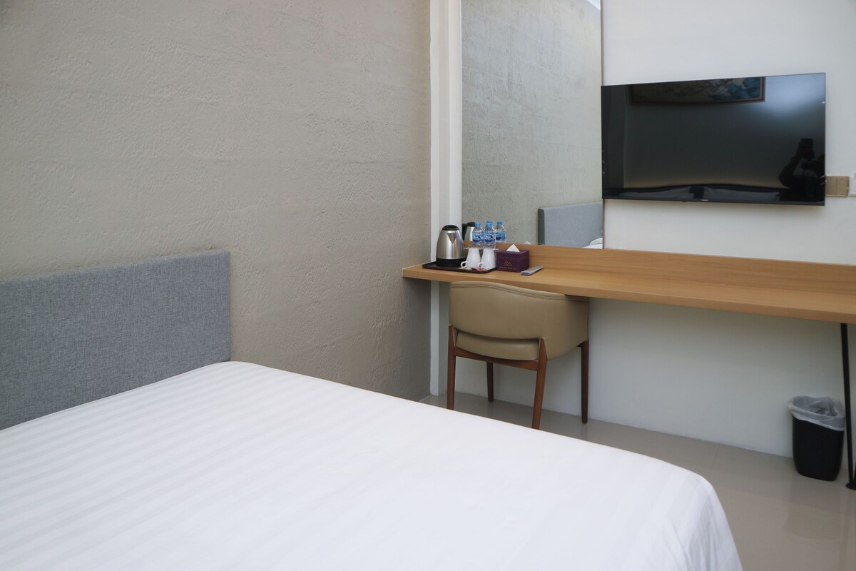 A hotel room with a bed, a desk with a chair, a television, and a trash bin.