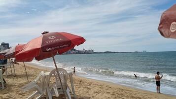 Uma praia com guarda-sóis e cadeiras, vista para o mar e um horizonte urbano distante.