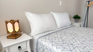 A neatly made bed with white pillows and a patterned bedspread, a bedside table with a lamp, and a small potted plant on a nightstand. - ASA Pe na Areia 3QT Beto Carrero Gravata (Navegantes)