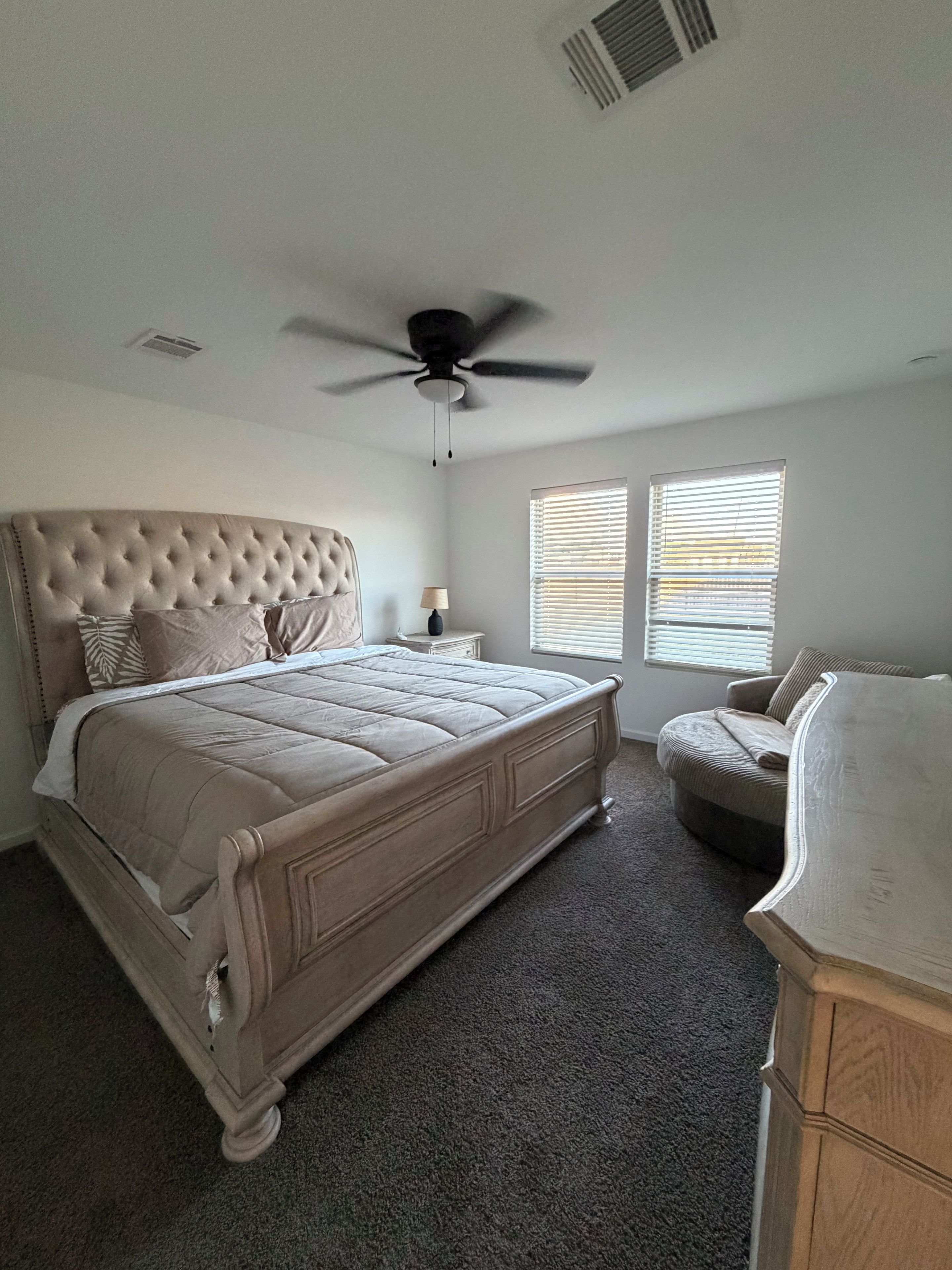 A bedroom with a large bed, a ceiling fan, two windows with blinds, and a nightstand.
