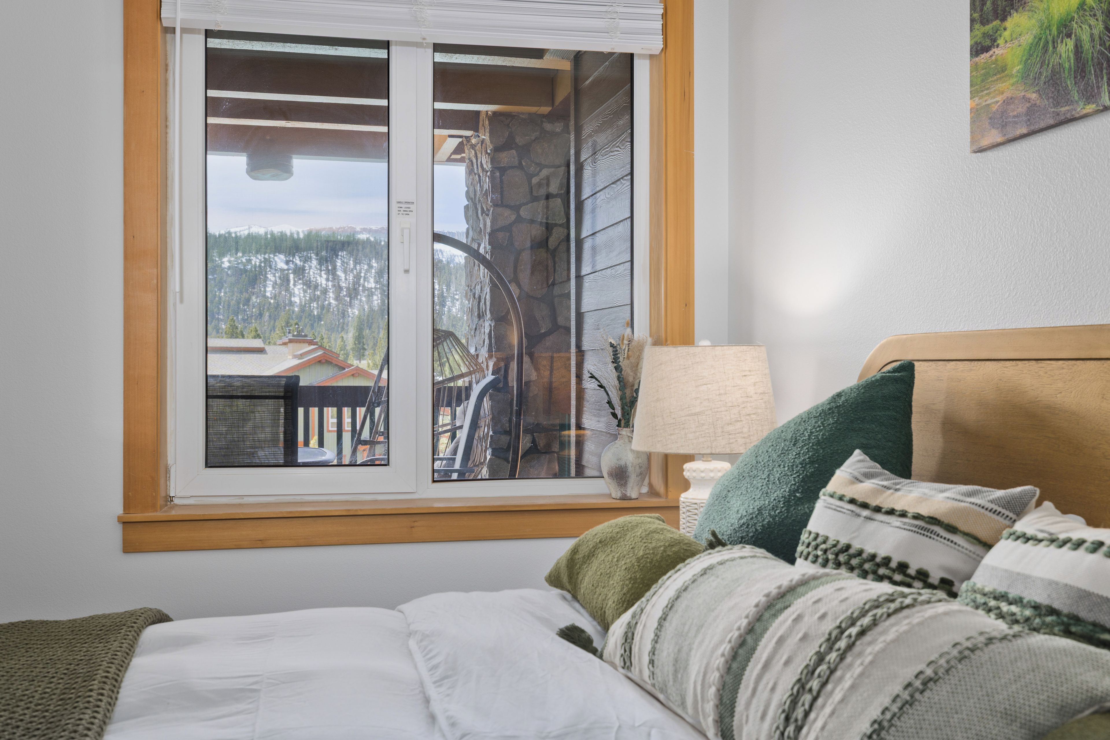 A bedroom with a bed, pillows, a nightstand, a lamp, and a window with a view of a cabin and forest.