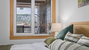 A bedroom with a bed, pillows, a nightstand, a lamp, and a window with a view of a cabin and forest.