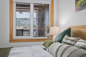 A bedroom with a bed, pillows, a nightstand, a lamp, and a window with a view of a cabin and forest.