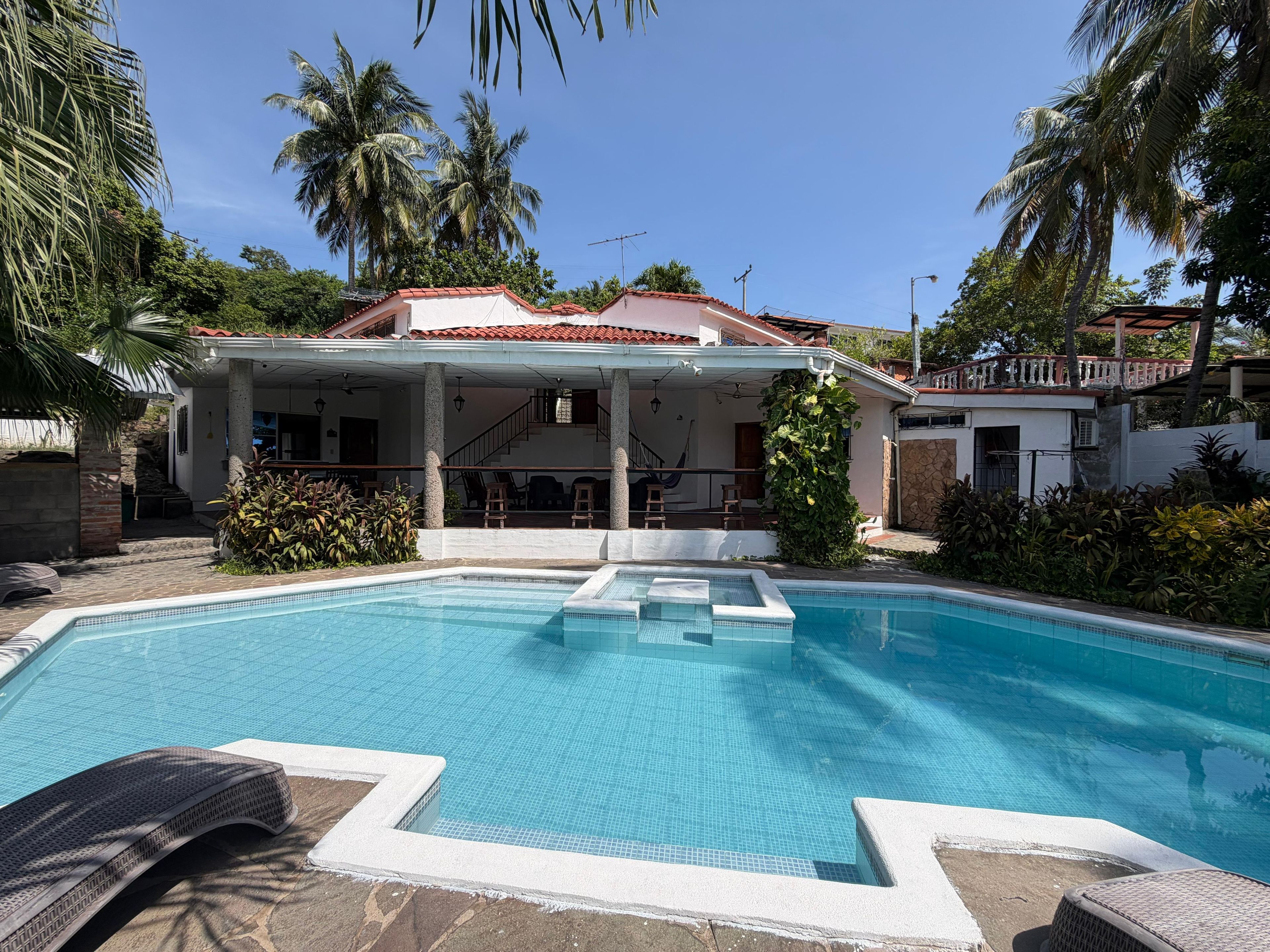 A swimming pool with a covered patio area, palm trees, and a house with a red roof.