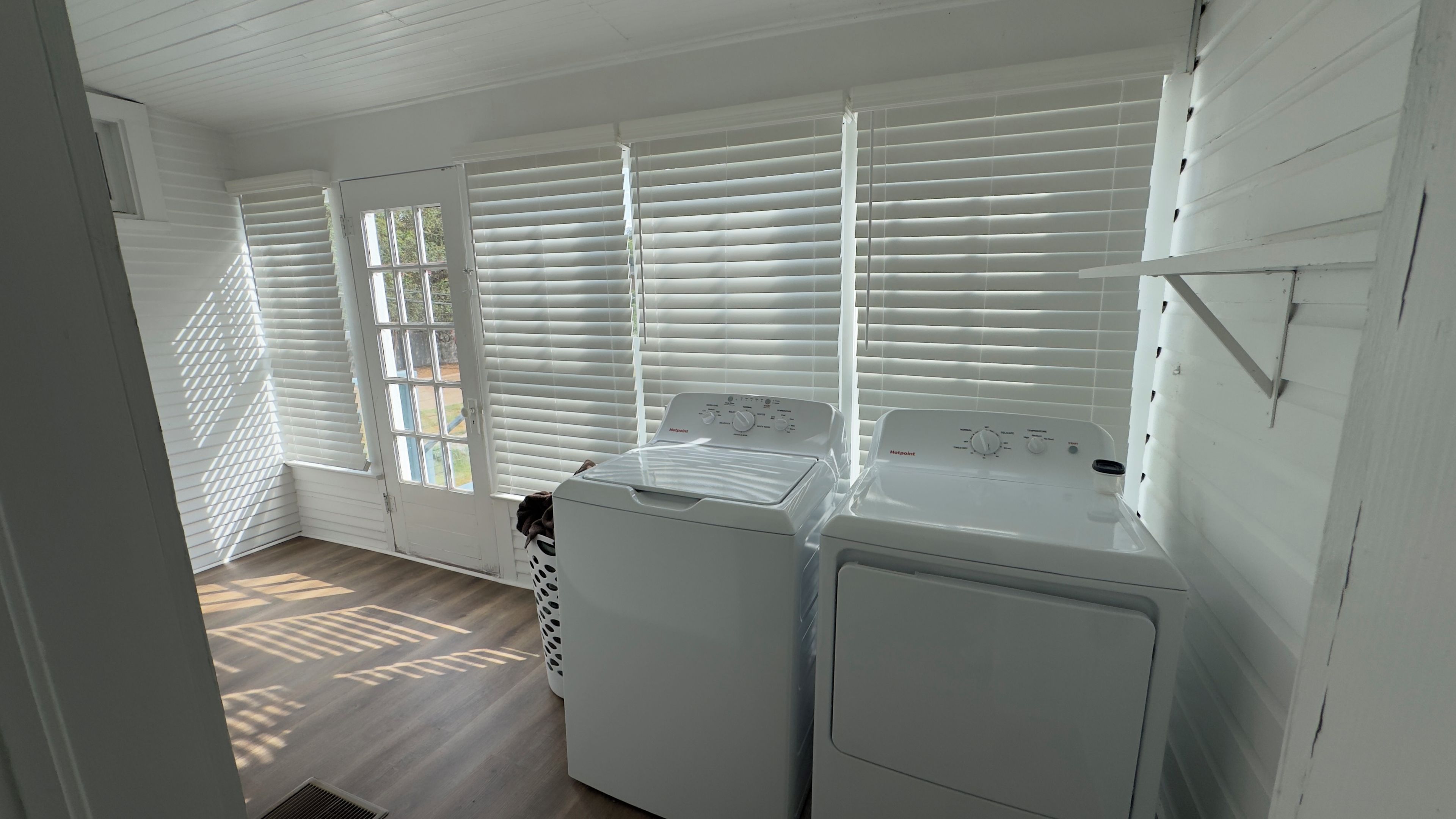 A laundry room with a front-loading washer and dryer, white blinds, and a wooden floor.