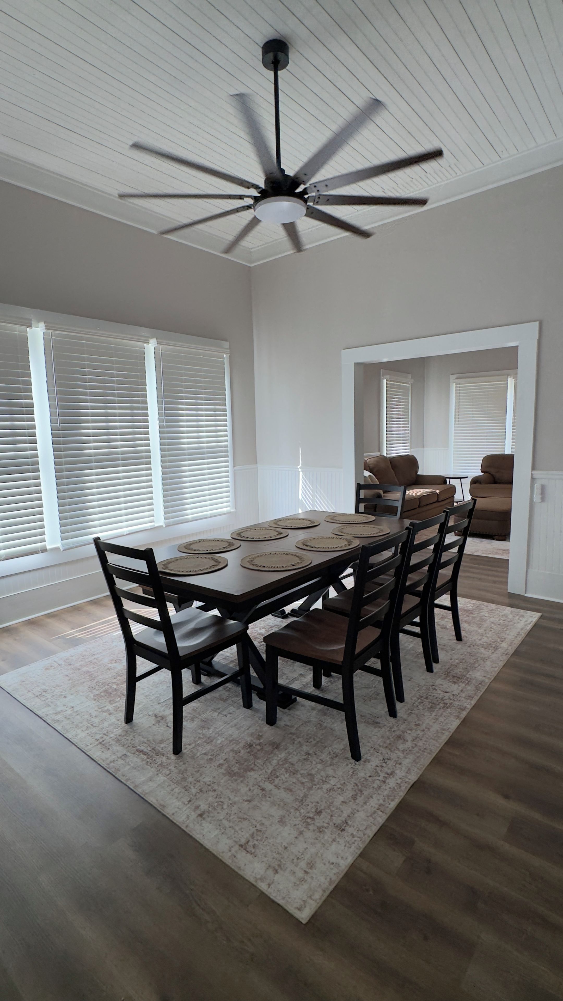 A dining area with a ceiling fan, a dining table set for six, and a view of the living room through an open doorway.