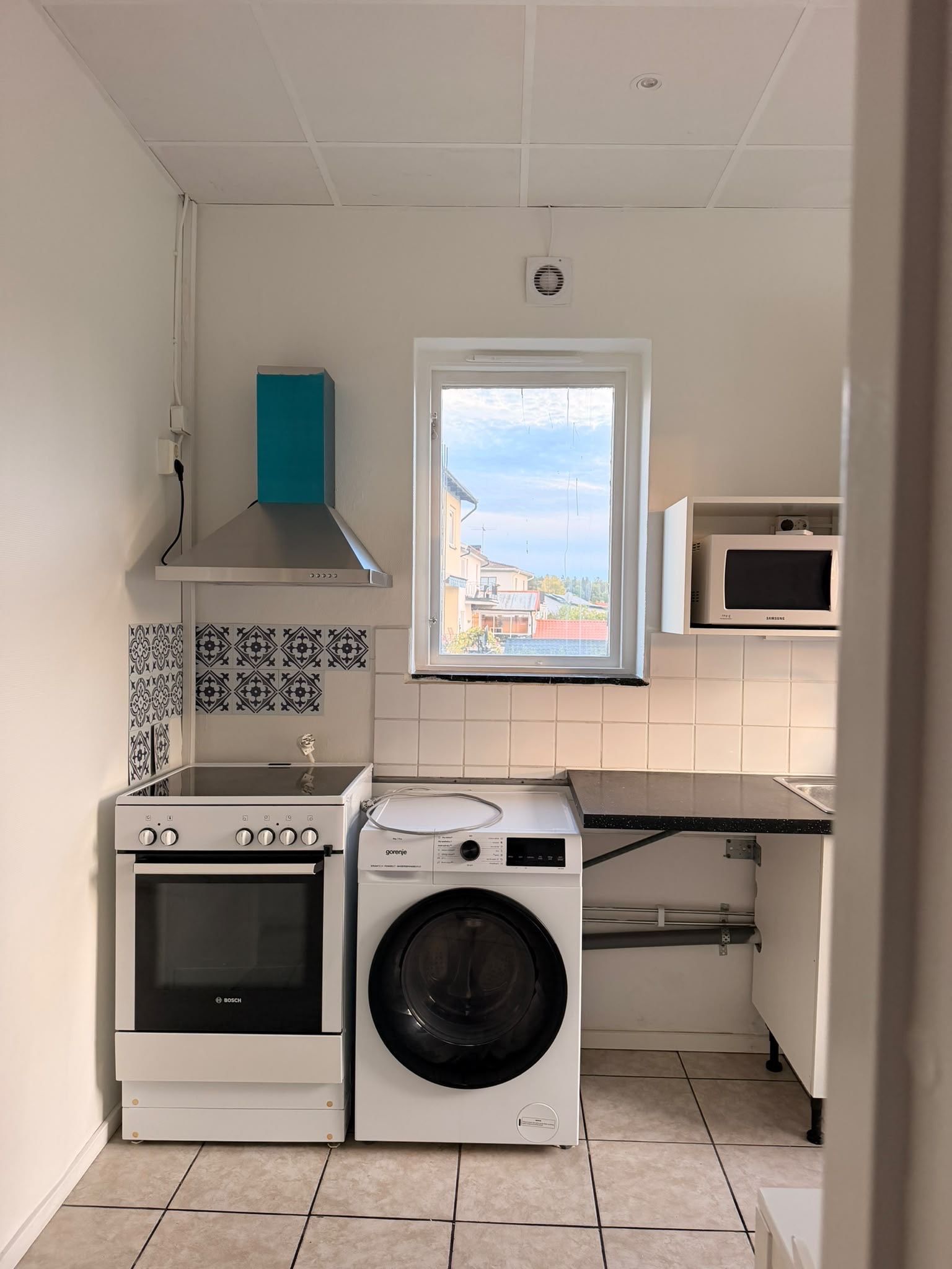 Una cucina compatta con forno Bosch, lavatrice, microonde e una finestra vista edifici.