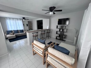 A modern living room with a sofa set, a dining area with bar stools, a kitchen in the background, and a ceiling fan. - Nice and accessible apartment in San Juan area, very walkable distance to malls. (San Juan)