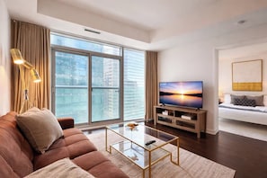 Een moderne woonkamer met een groot raam, een flatscreen televisie, een bruine bank en een glazen salontafel.