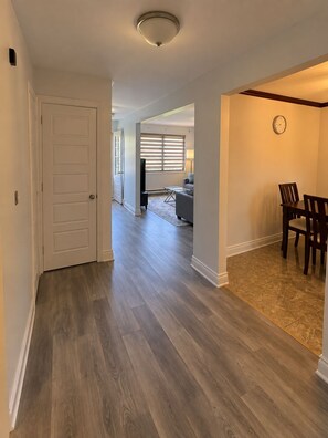 Un couloir avec une porte donnant sur un salon, un coin repas avec une table et des chaises, et une horloge au mur.