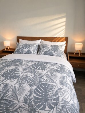 A bed with a tropical leaf pattern bedspread, flanked by two bedside tables with lamps, in a room with a plain wall and a wooden floor. - Cozy Stay • Large Bedrooms • Smart TV • Parking (Montréal)