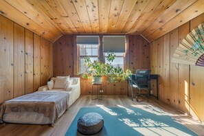 Quarto com paredes e teto de madeira, uma cama, uma escrivaninha com cadeira, uma janela com persianas e um vaso de plantas.