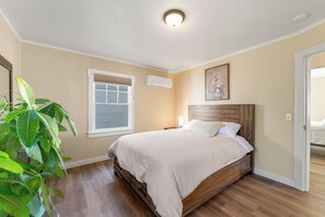 A bedroom with a bed, a window with blinds, an air conditioning unit, a painting on the wall, and a plant. - Bright Mt. Shasta Cottage - Hot Tub, Patio & Coffee Bar (Mount Shasta)
