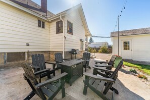 Een terras met Adirondack-stoelen en een barbecue, tegen een huis met een schuin dak en schoorsteen.