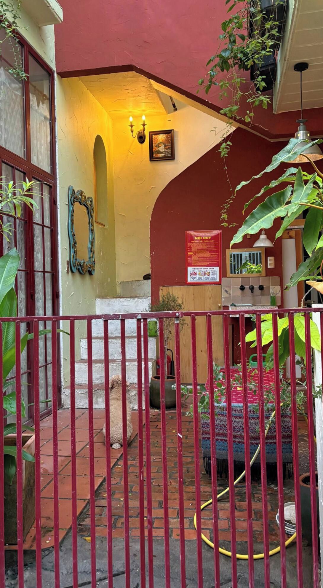 Un patio con una reja metálica roja, suelo de baldosas y pared amarilla con un cuadro enmarcado.