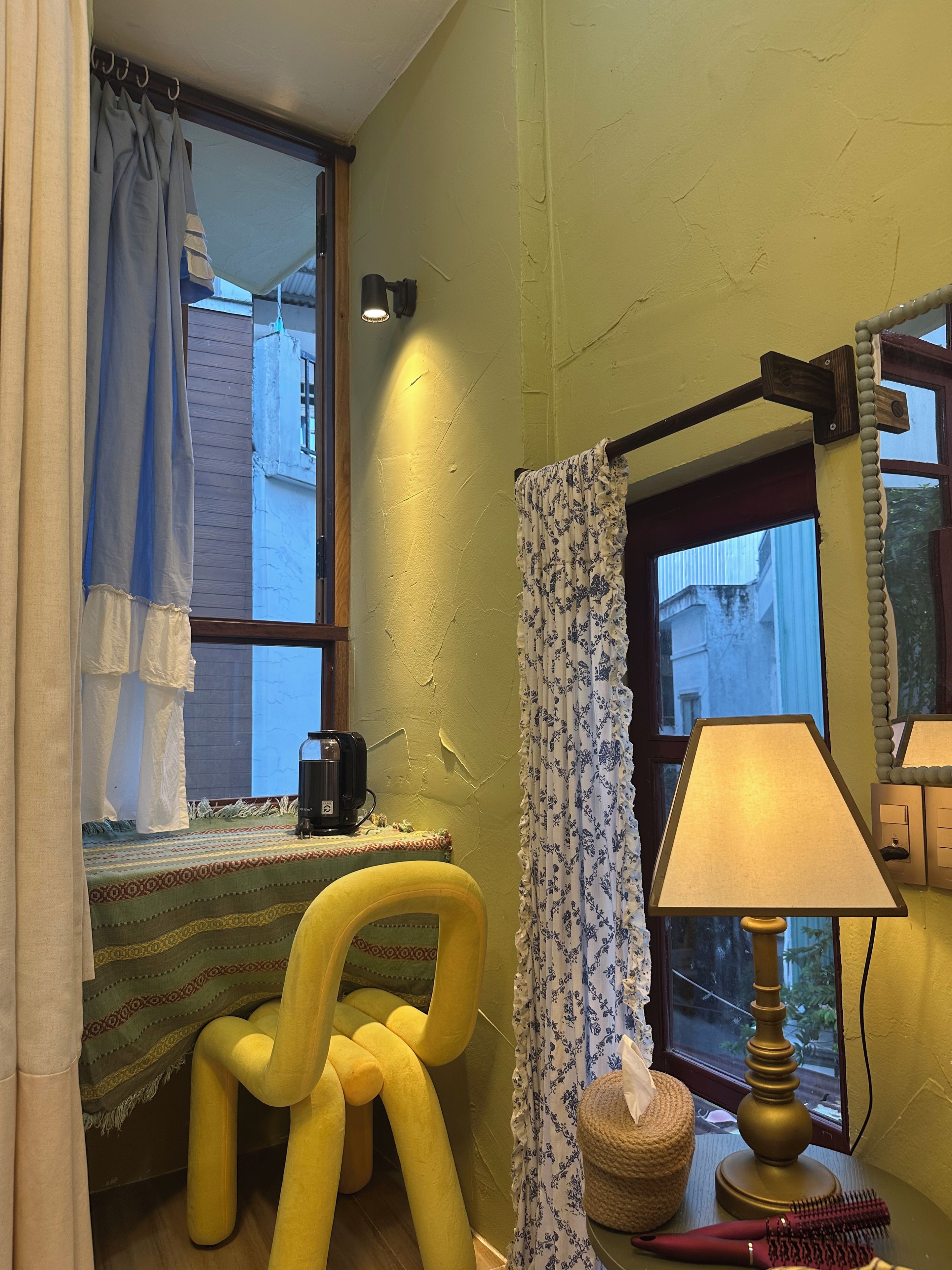Una habitación con ventana, una pequeña mesa con lámpara, una silla y un objeto decorativo sobre la mesa.