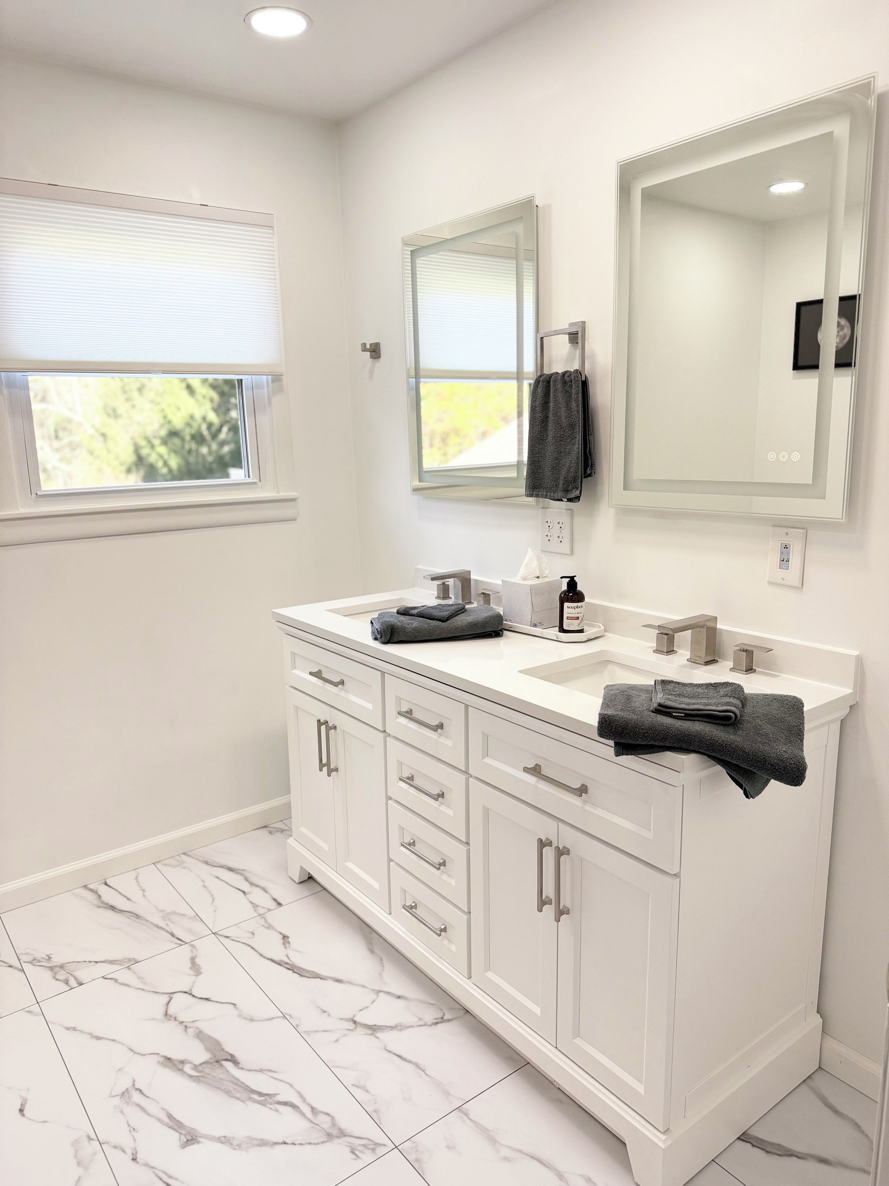 A bathroom with a white vanity, two sinks, a large mirror, and a window with blinds.