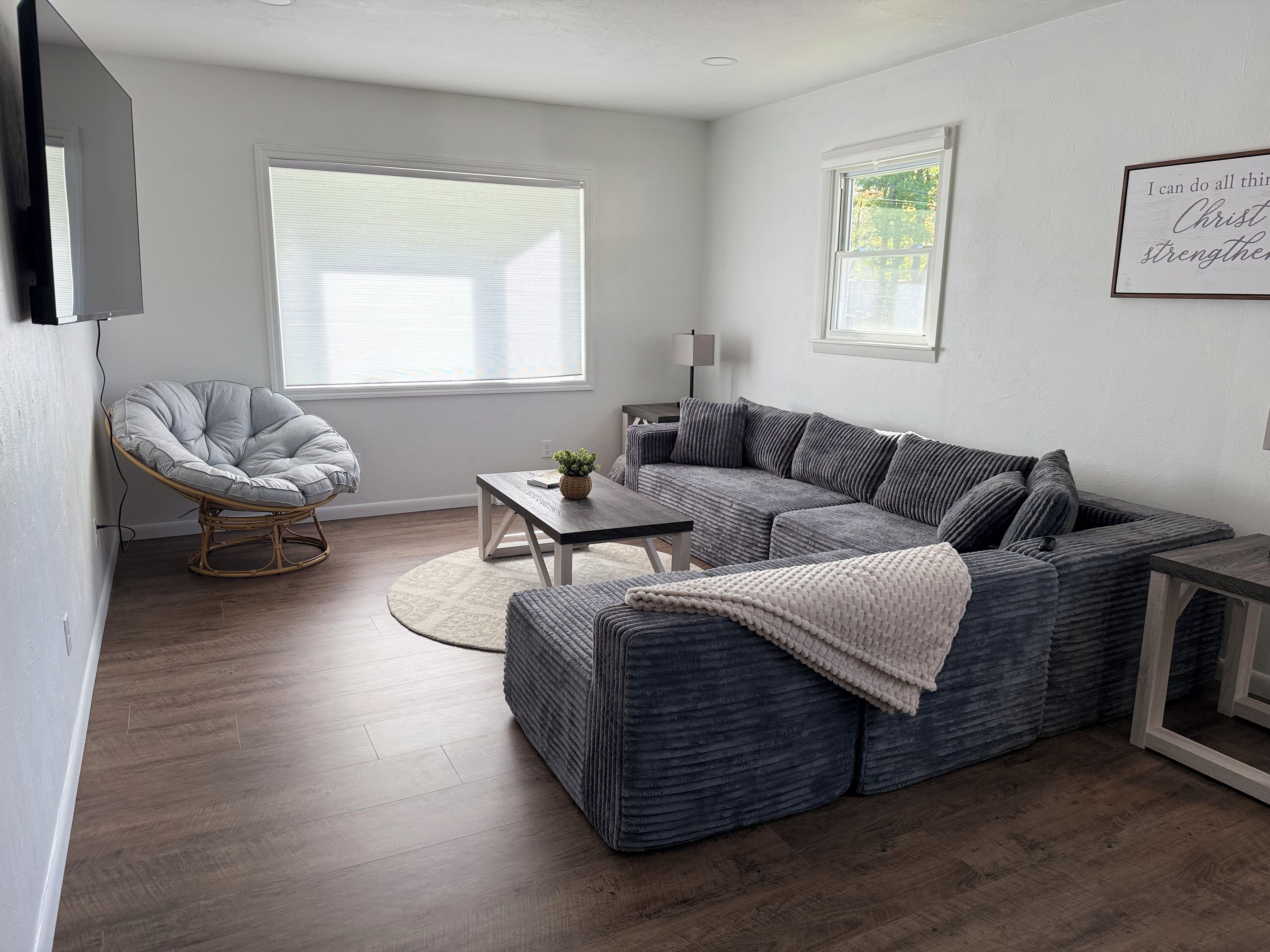 Ein modernes Wohnzimmer mit einem grauen Ecksofa, einem runden Sessel, einem kleinen Tisch und einem großen Fenster.