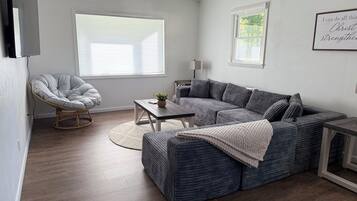 Ein modernes Wohnzimmer mit einem grauen Ecksofa, einem runden Sessel, einem kleinen Tisch und einem großen Fenster.
