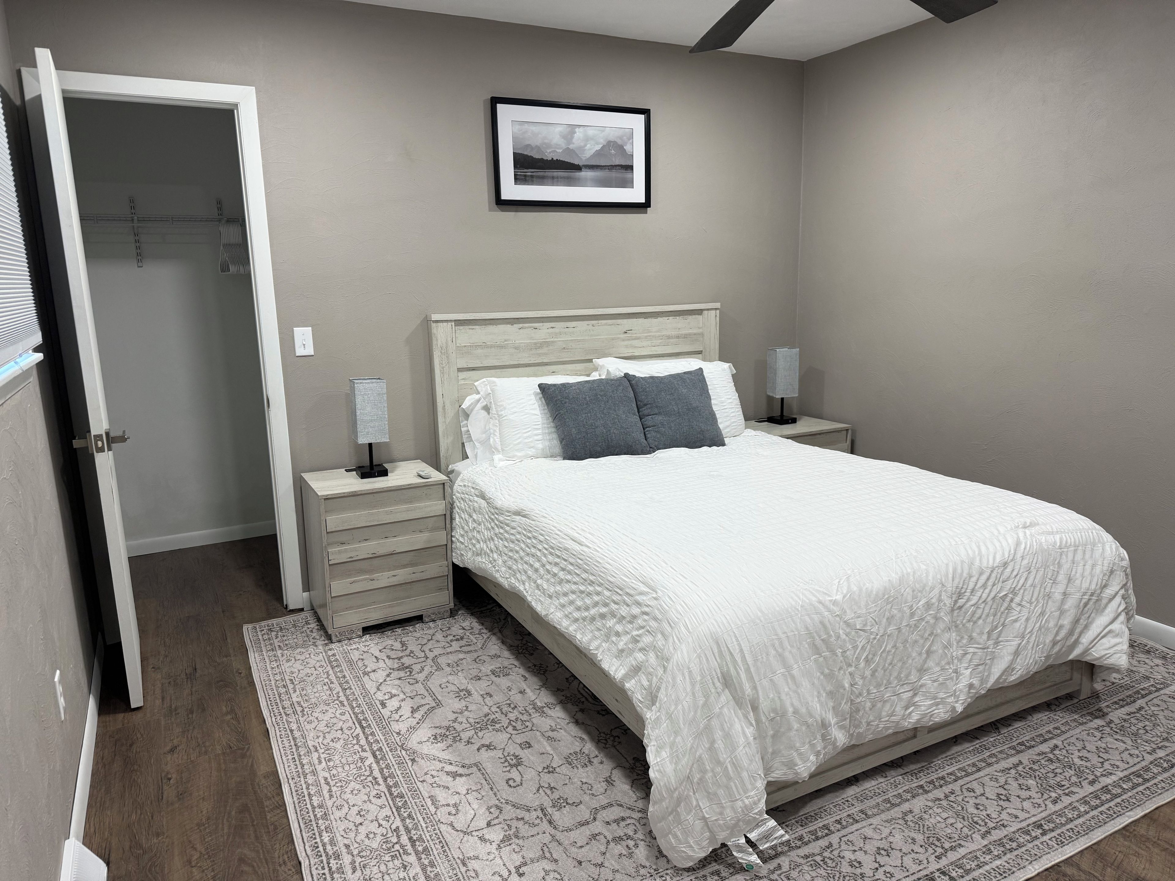 A bedroom with a bed, bedside tables, a nightstand, a rug, and a framed picture on the wall.